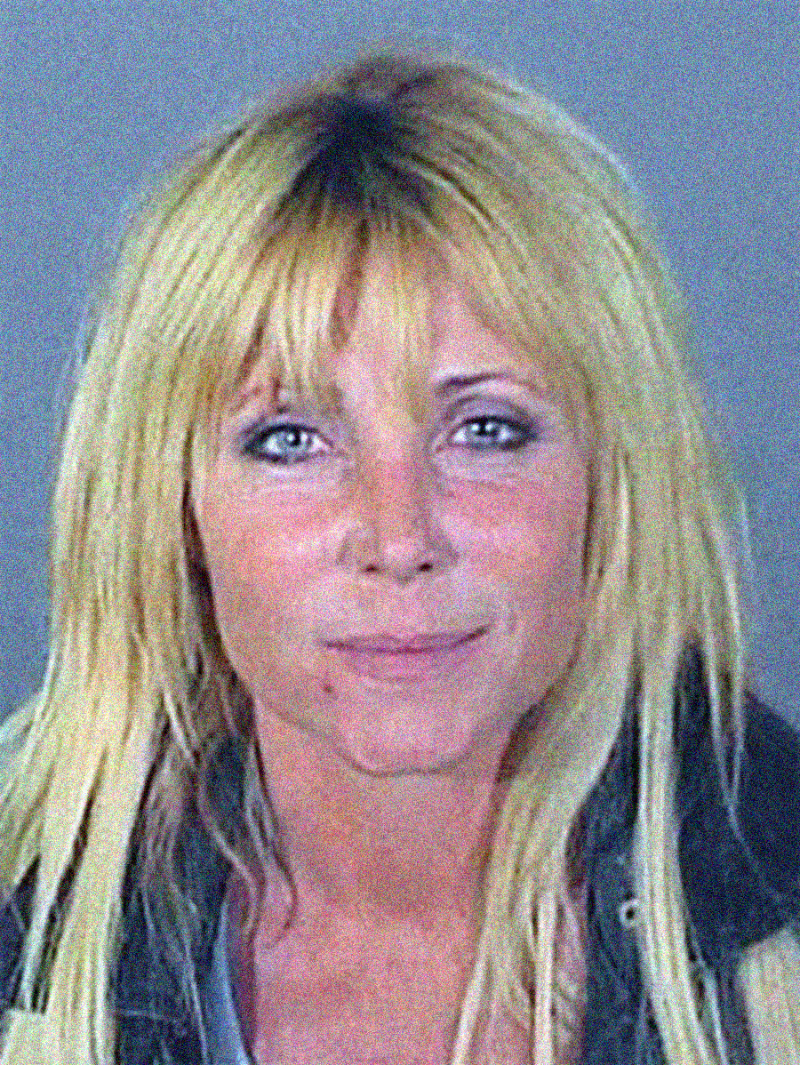 A woman with long, messy blonde hair and bangs looks at the camera with a slight smile, wearing a dark jacket over a light top, in front of a plain gray background.