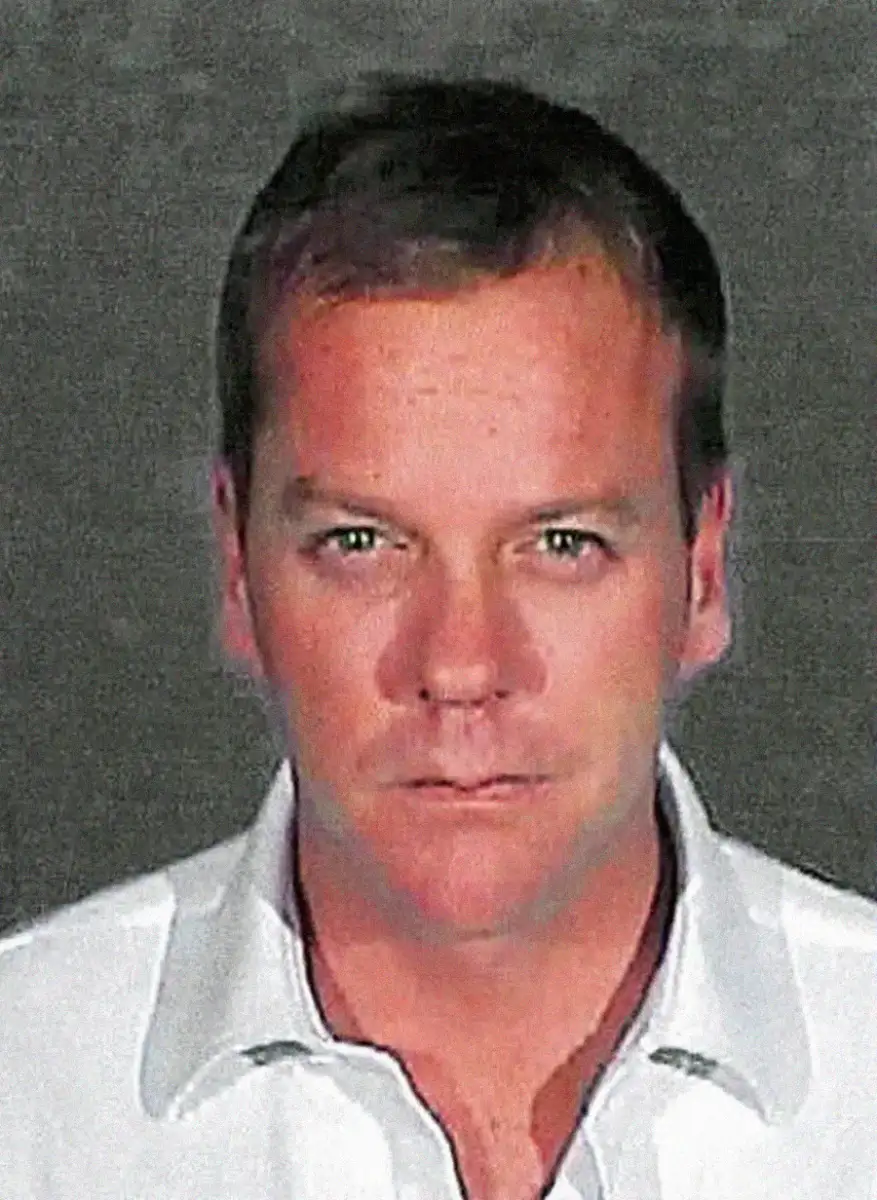A man with short hair wearing a white collared shirt looks straight at the camera against a plain, dark background.