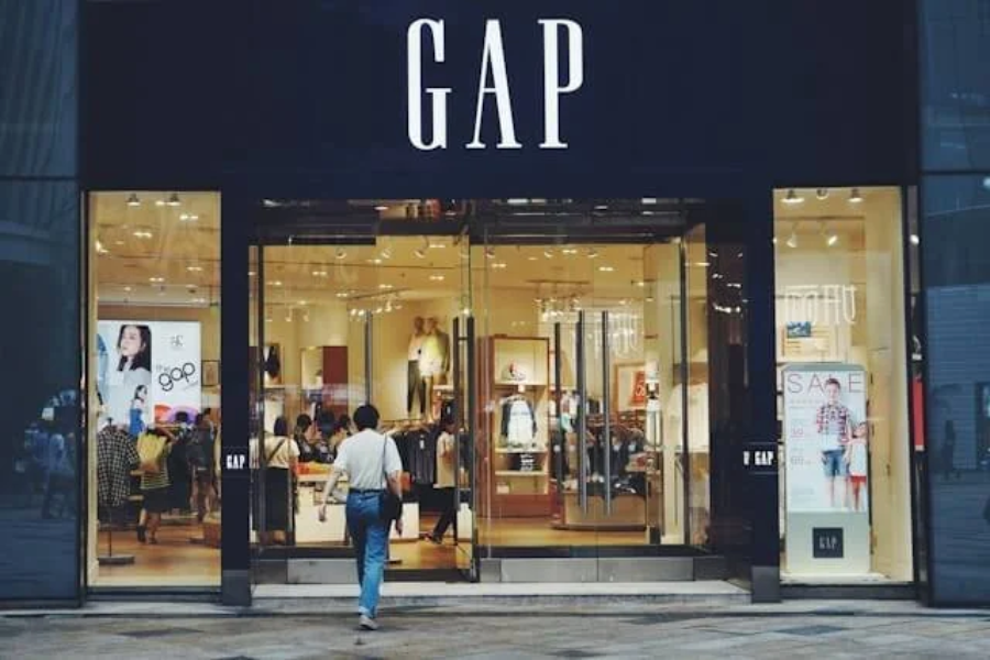 A person enters a well-lit GAP clothing store with large glass doors. The store's black sign with bold white GAP letters is visible above the entrance. Mannequins and clothes are displayed inside.