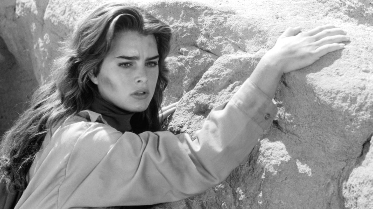 A woman with long hair and a concerned expression grips a rocky surface with one hand, looking into the distance. The image is in black and white.