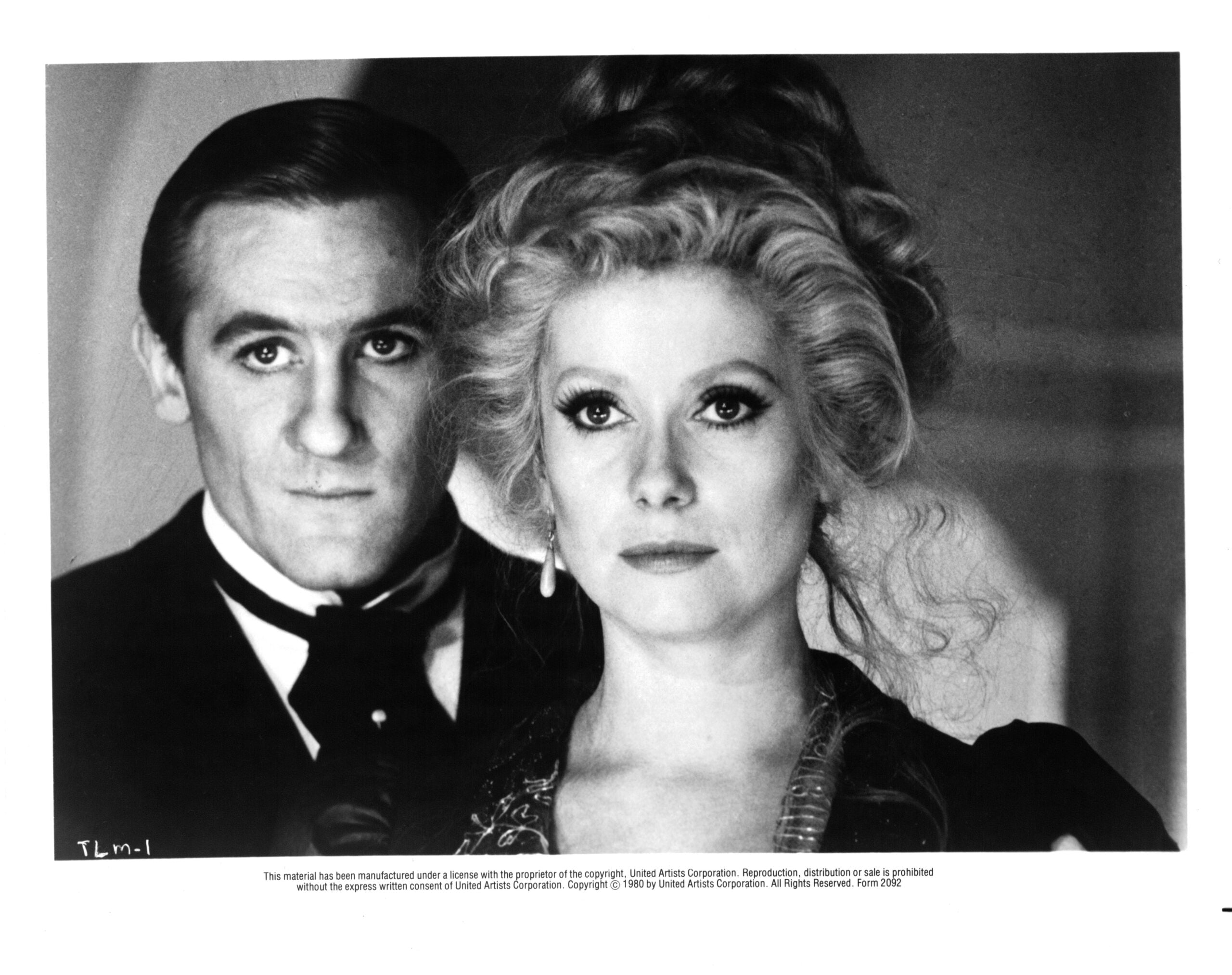 A man in a dark suit stands beside a woman with styled blonde hair in an ornate dress. They both face forward, appearing serious, in a black-and-white portrait with a vintage atmosphere.