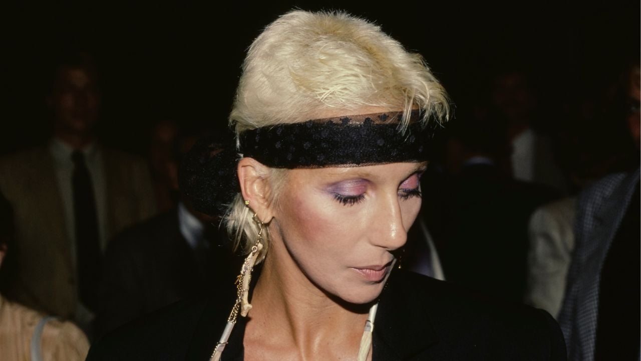 A person with short, bleached blonde hair and a black headband looks down. They wear dramatic makeup, a dangling earring, and a black outfit. There are blurred people in the dark background.