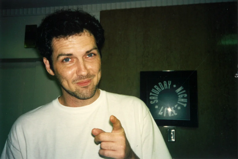 A man with short dark hair wearing a white T-shirt smiles and points at the camera. Behind him is a framed "Saturday Night Live" sign on a wall.