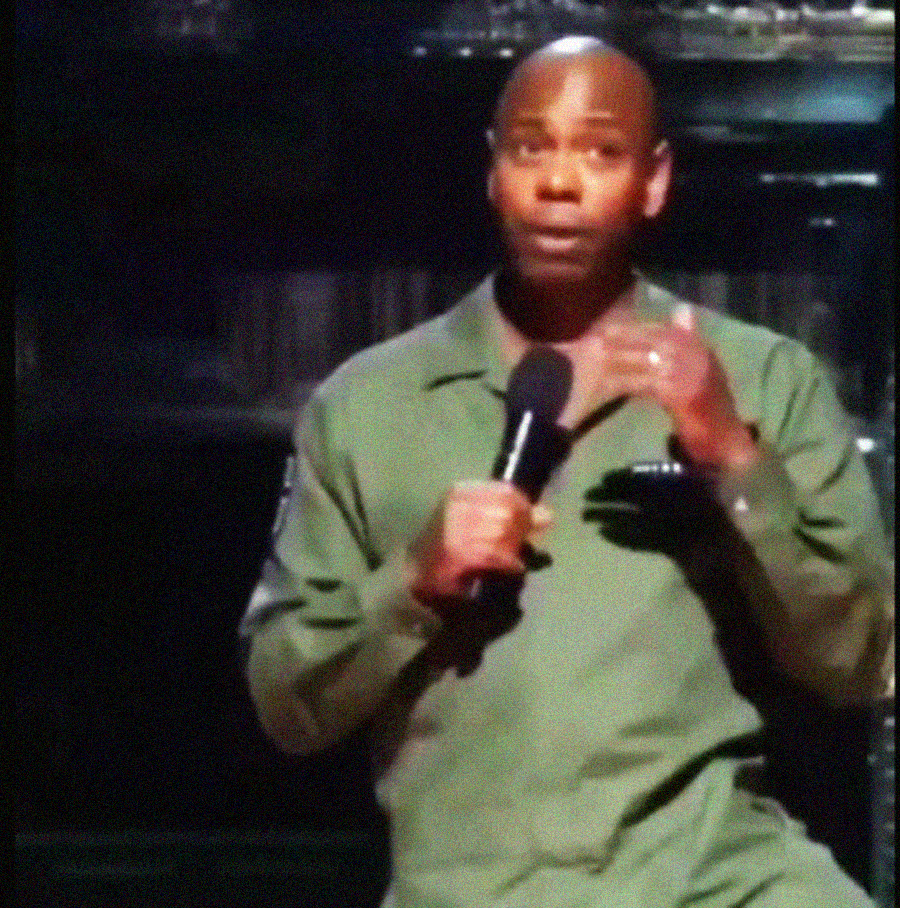 A man in a green jacket holds a microphone and gestures with his hand while performing on stage, with a dark background behind him.