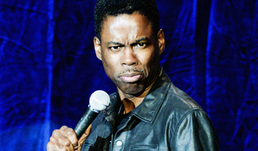 A comedian wearing a black leather jacket holds a microphone and makes a humorous, expressive face while performing on stage in front of a blue curtain.