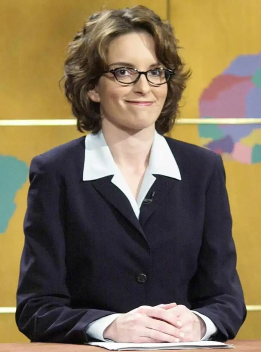 A woman with short brown hair, glasses, and a dark suit sits at a news desk, smiling slightly. The background is yellow with abstract colored shapes and lines.