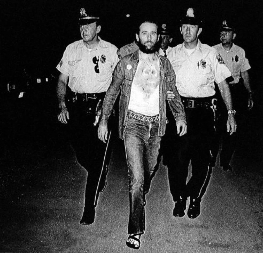 A bearded man in casual clothes is escorted by three uniformed police officers at night. The officers hold his arms as they walk, and the scene appears tense and serious.