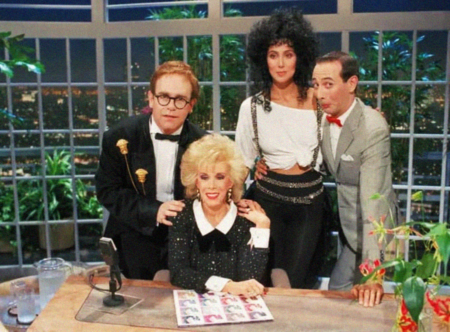 Four people pose on a late-night TV show set. A blond woman in a sparkly black outfit sits at a desk, while three others—two men in suits and a woman with dark curly hair—stand behind her, all smiling.