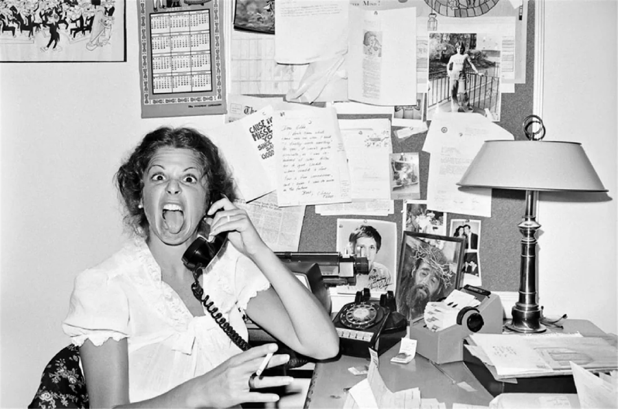A woman with an exaggerated shocked expression talks on a rotary phone at a cluttered desk, covered with papers, photos, a lamp, and a bulletin board full of notes and pictures.