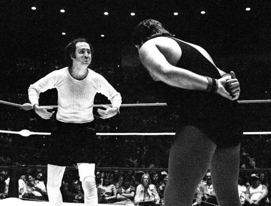A black-and-white photo of two men in a wrestling ring. One man, in a light shirt and dark shorts, gestures with his hands. The other man, in a singlet, stands with hands behind his back. An audience is visible in the background.