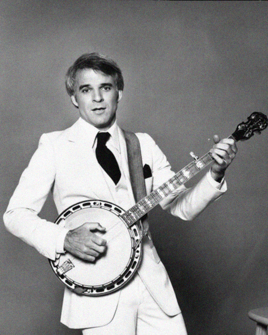 A man in a white suit and black tie plays a banjo against a plain background, looking confidently at the camera.