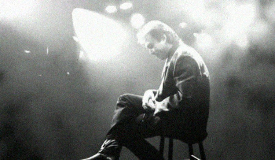 A person in a suit sits on a stool, head bowed, under dramatic stage lighting with a smoky, high-contrast atmosphere.