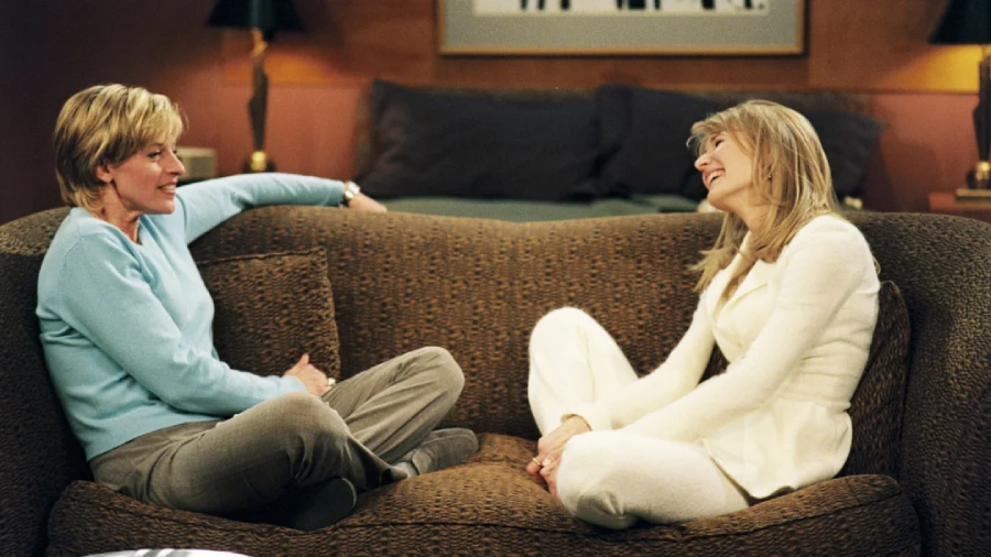 Two women sit facing each other on a brown sofa, smiling and talking, in a cozy room with warm lighting and cushions in the background.