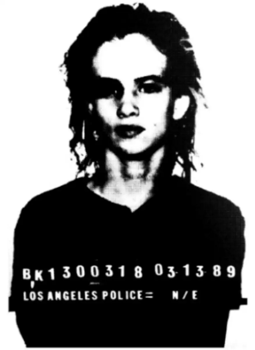 Black and white mugshot of a person with medium-length hair wearing a dark shirt, looking straight at the camera. Text below shows “LOS ANGELES POLICE,” a booking number, and the date “03-13-89.”