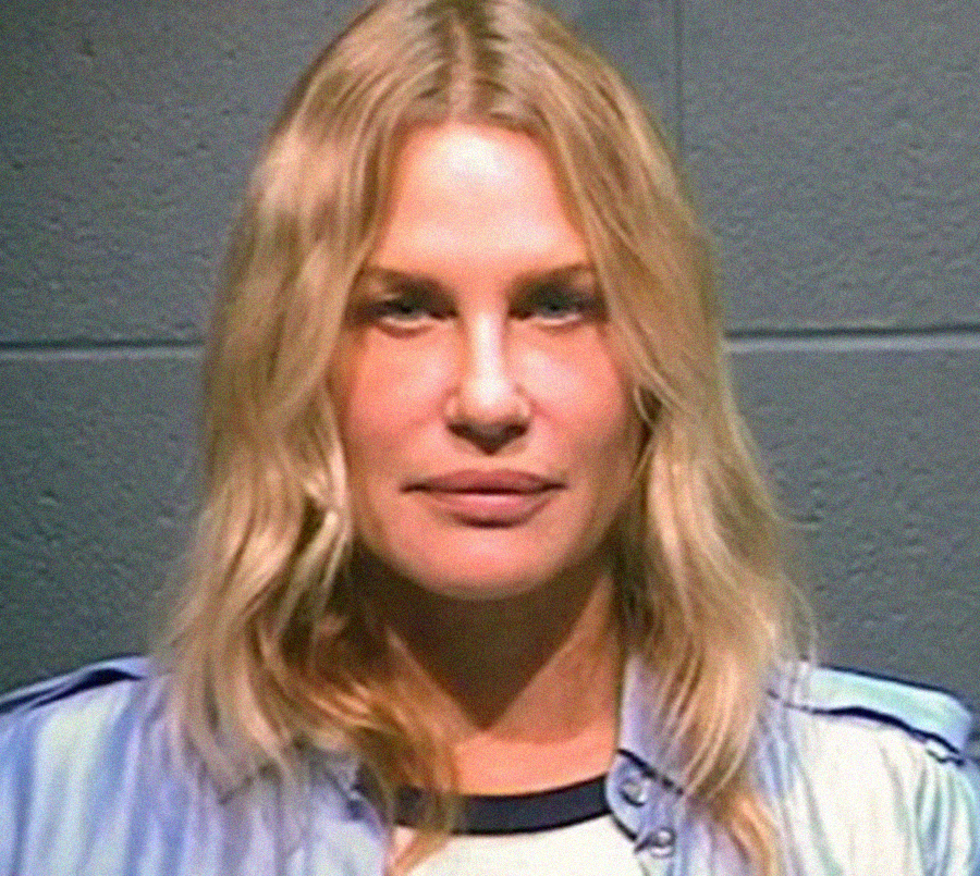 A woman with long blonde hair and a neutral expression stands against a gray cinder block wall, wearing a light blue shirt over a white top.