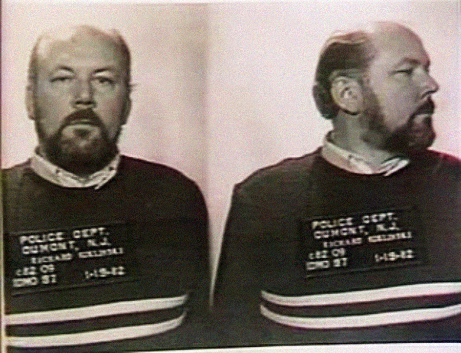 Mugshot of a man with a beard and mustache, shown in both front and side profiles. He is wearing a dark sweater with white and red stripes and a police identification placard.