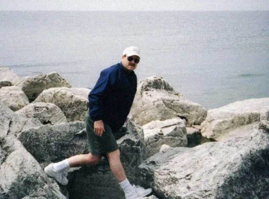 A man in a blue jacket, shorts, white sneakers, and a cap stands on large rocks by a calm body of water under a cloudy sky.