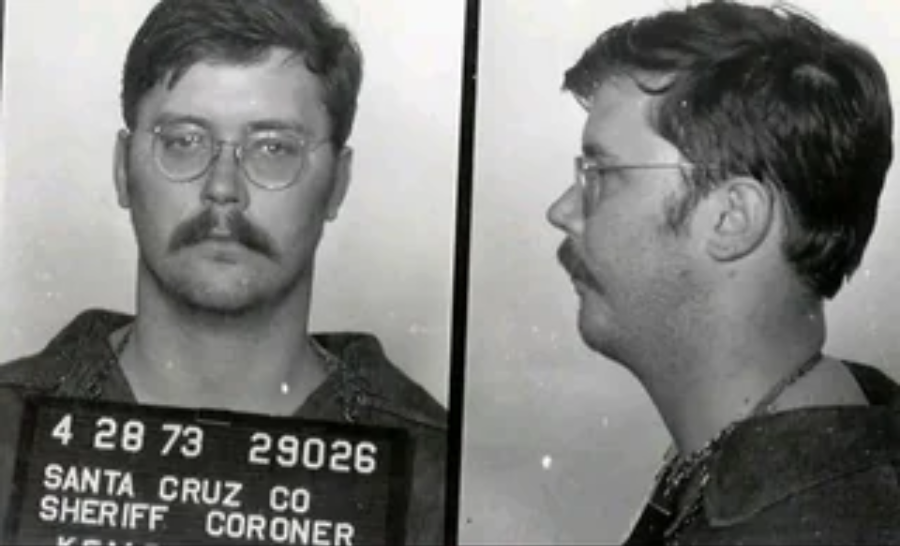 Black and white mugshot with front and side views of a man wearing glasses and a mustache, holding a sign with details including "Santa Cruz Co Sheriff Coroner" and the date "4 28 73.