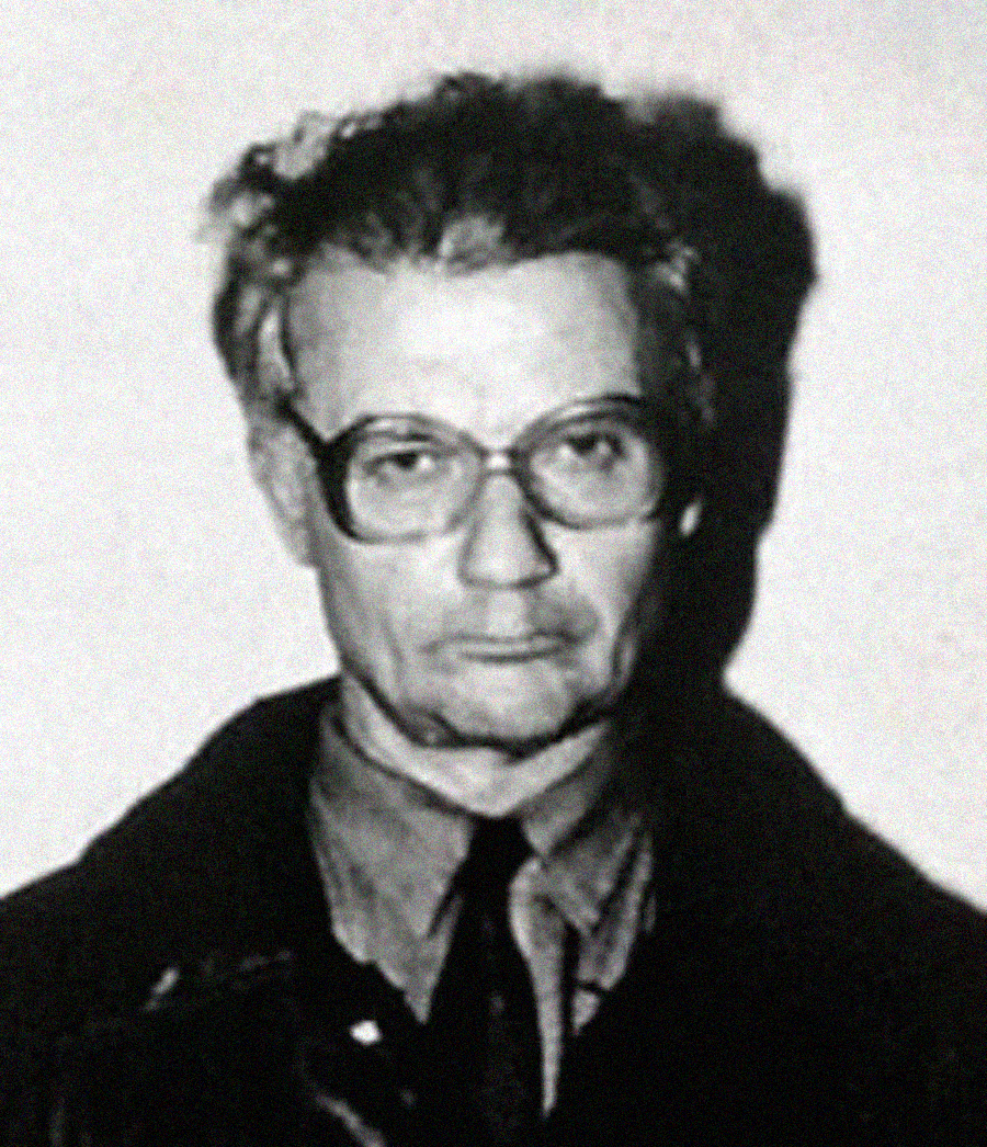 A black and white portrait of a middle-aged person with short hair, glasses, and a serious expression, wearing a collared shirt, tie, and jacket, facing forward against a plain background.