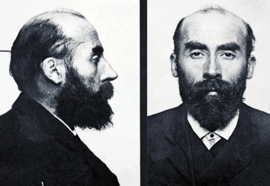 Black and white mugshot photo showing a man with a full beard. The left side displays his profile, while the right side shows him facing forward with a serious expression, wearing a suit and collared shirt.