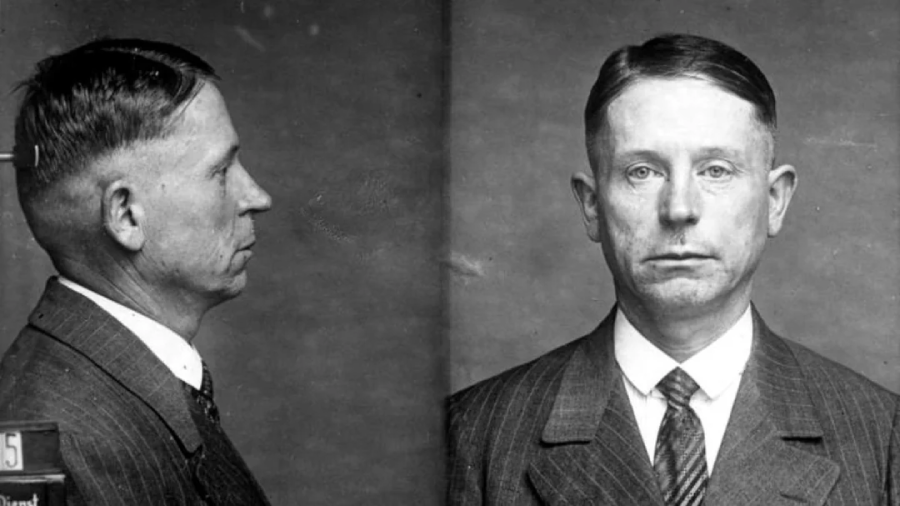 Black-and-white mugshot showing a man in a suit and tie, with one image in profile and the other facing forward. The man has short, neatly combed hair and a serious expression.