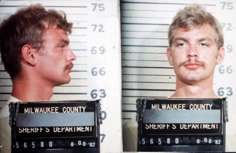 A mugshot showing a young man with light hair and a mustache, facing left in one photo and forward in another. He is standing in front of a height chart, holding a Milwaukee County Sheriff’s Department sign dated 8-08-82.