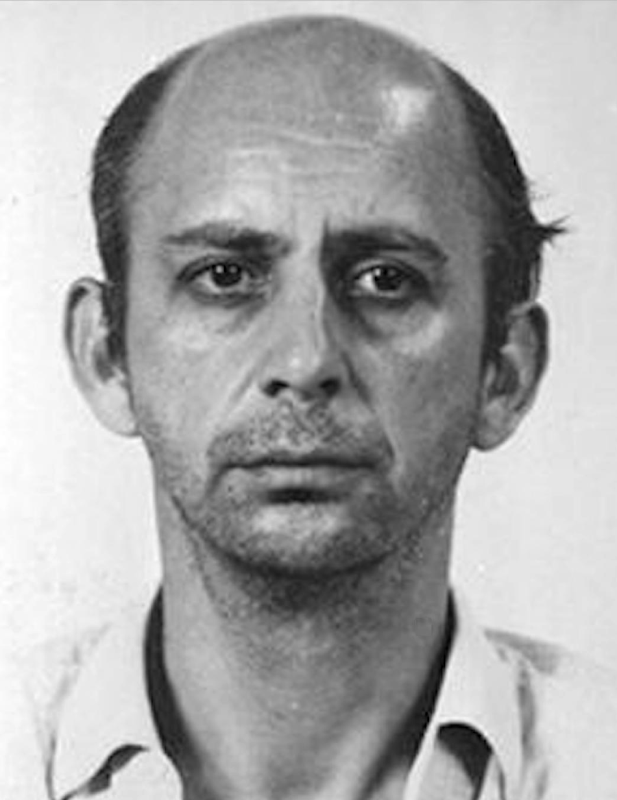 Black and white portrait of a middle-aged man with a receding hairline, thinning hair, and a serious expression. He is wearing a light-colored collared shirt and is facing the camera against a plain background.