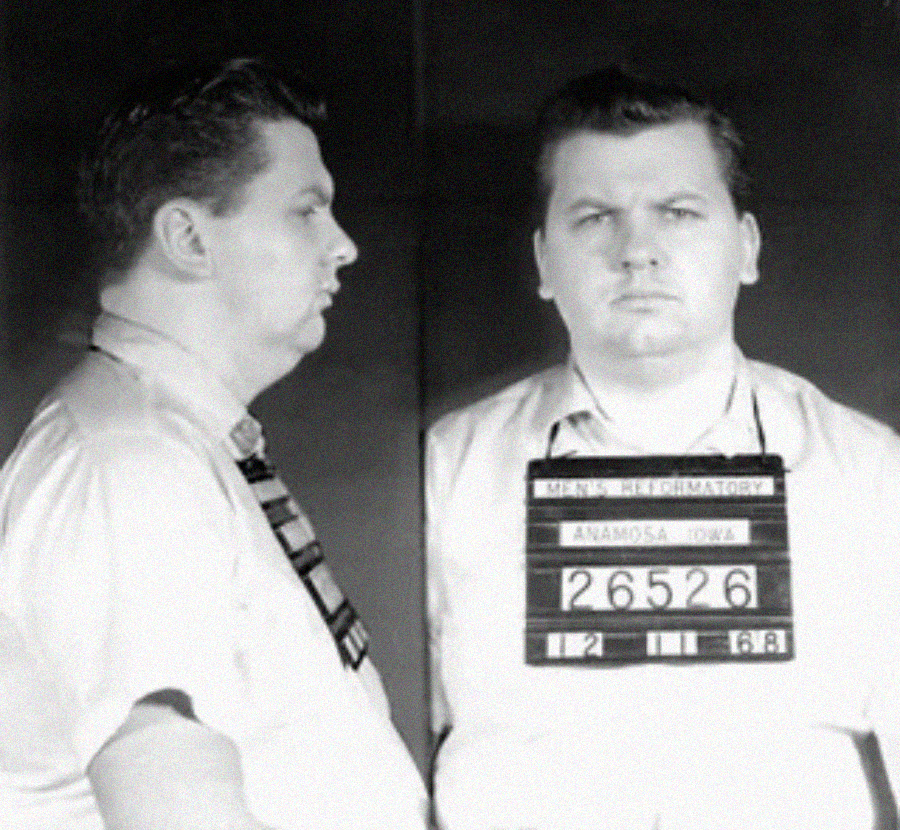Black and white mugshot of a man in a striped tie and collared shirt, shown in profile and front view, holding a sign that reads “MEN'S REFORMATORY ANAMOSA IOWA 26526 12 11 68.”