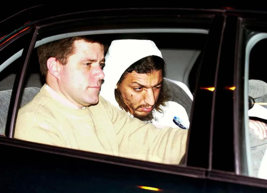 Two men sit in the front seats of a car at night. The man on the left wears a light sweater and has short hair, while the man on the right, in a white hoodie, appears disheveled and is looking down with a serious expression.