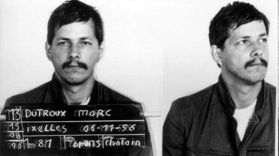 A man with short dark hair and a mustache is pictured in a black and white mugshot, shown facing forward and in profile. He wears a jacket, and a board with text displaying his name and details is held in front of him.