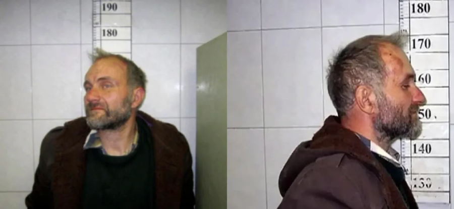 A man with a beard and disheveled hair stands against a height chart for two mugshot-style photos; one is a front view, the other a side profile. The background is tiled, and he wears a dark coat over a shirt.