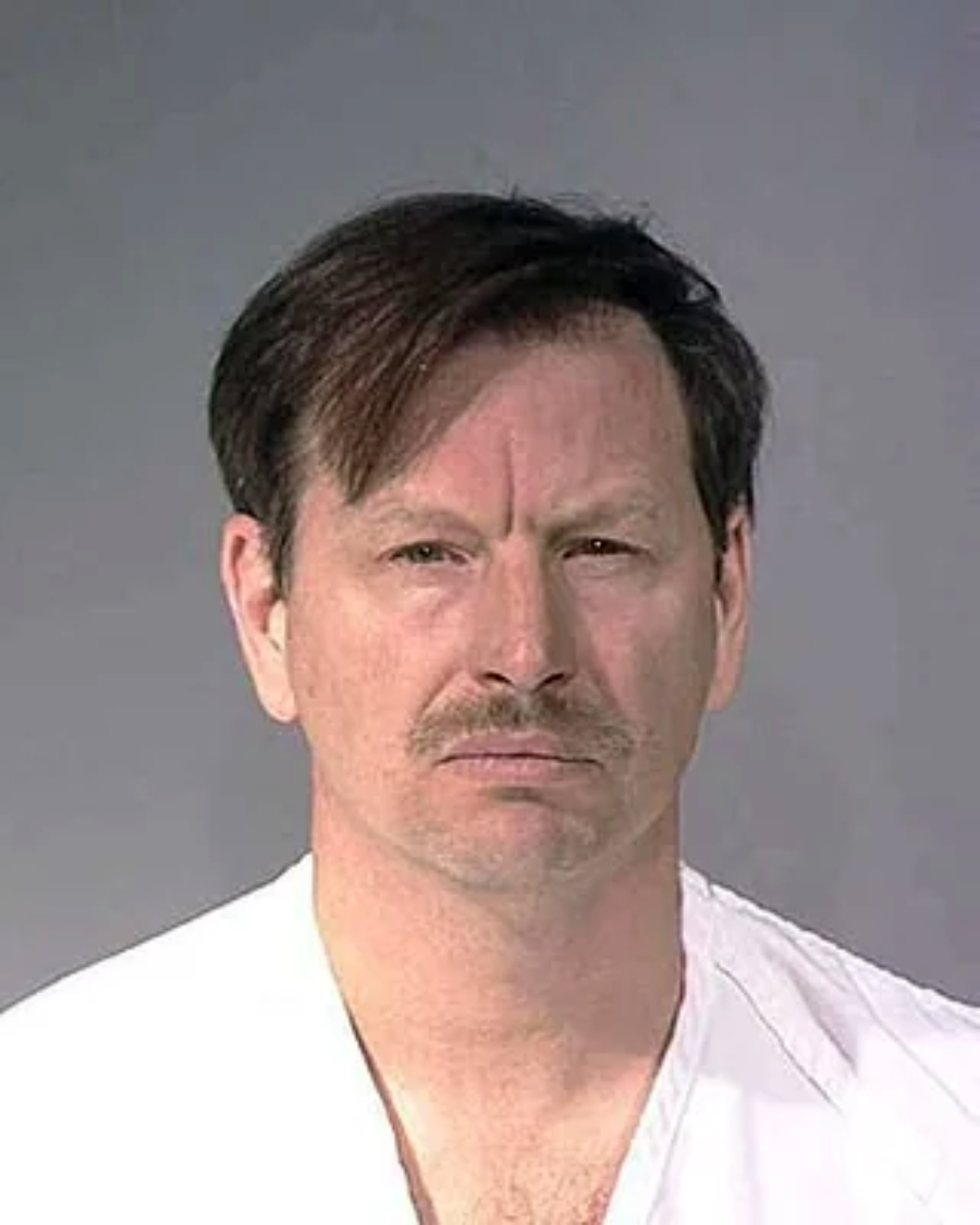 A middle-aged man with short brown hair and a mustache stands against a plain gray background, wearing a white shirt. He has a neutral expression and is looking straight at the camera.
