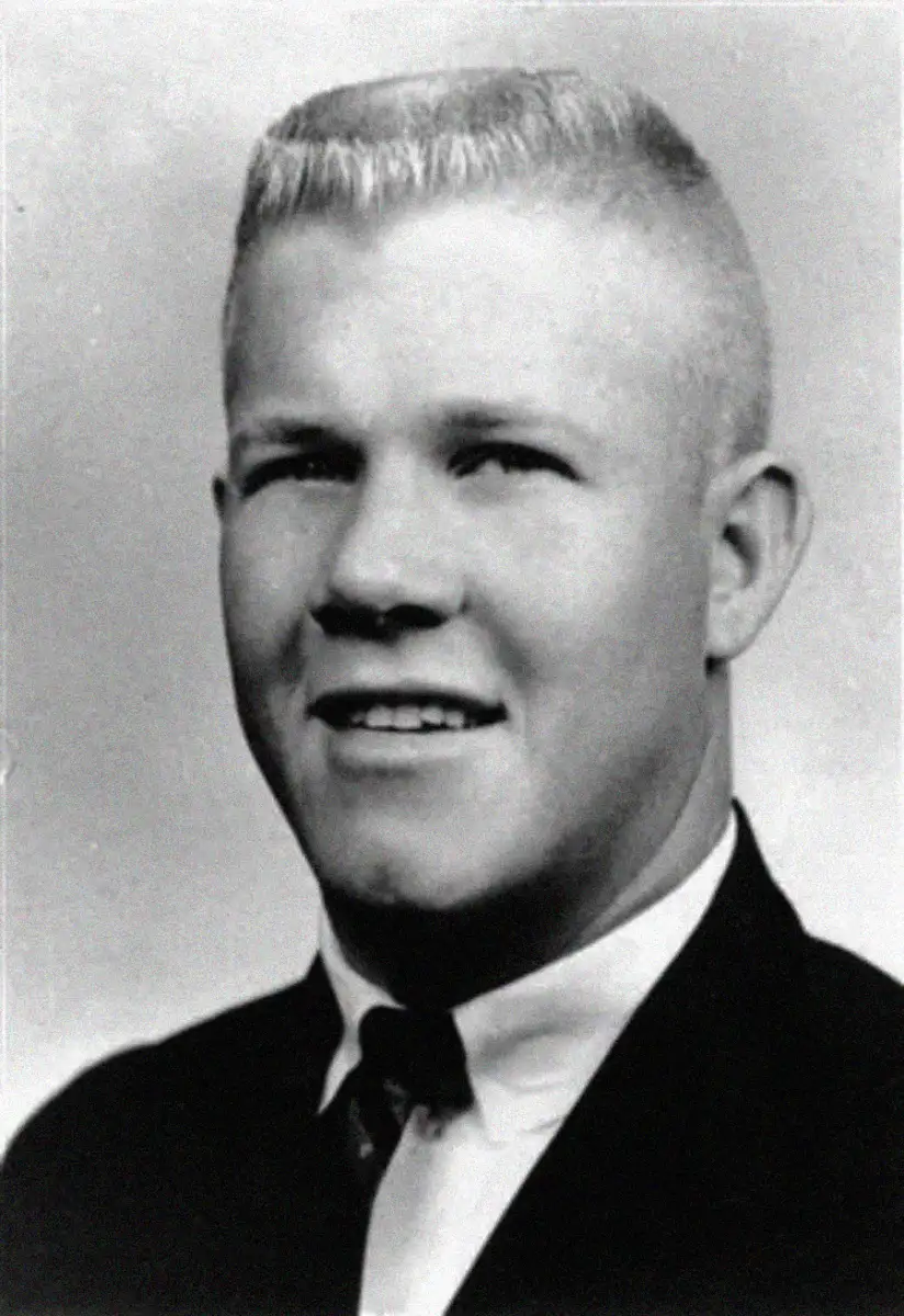 Black and white portrait of a young man with short, light-colored hair, wearing a suit jacket, dress shirt, and tie, looking slightly to the side and smiling faintly.