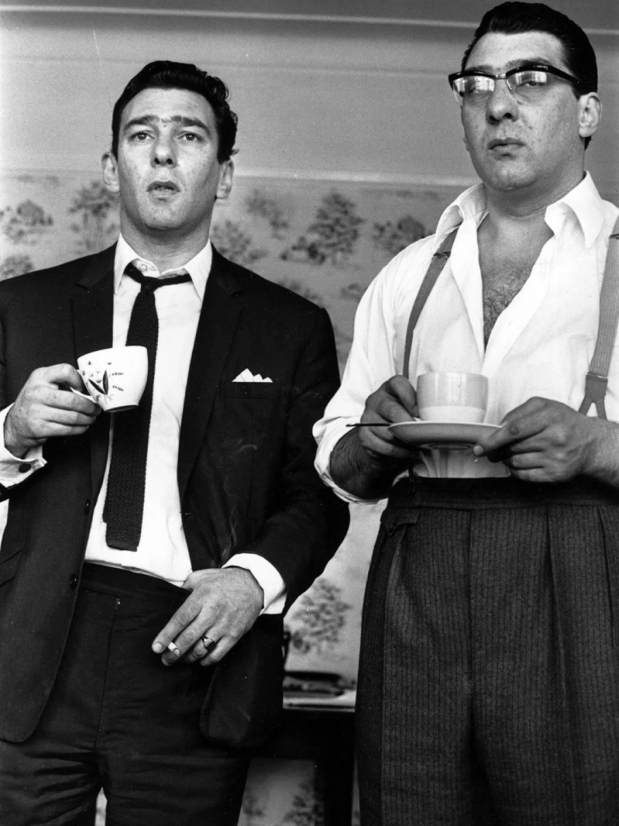 Two men stand indoors, both holding cups and saucers. One wears a suit and tie, the other wears glasses, a white shirt with rolled-up sleeves, suspenders, and high-waisted trousers. The background has patterned wallpaper.