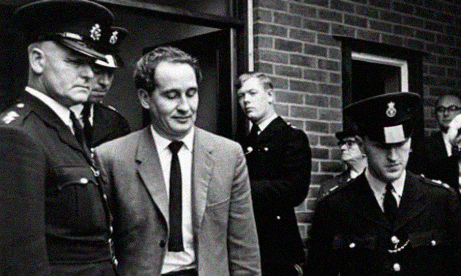 A man in a suit is flanked by police officers in uniform as he exits a brick building, with several people standing in the background. The scene appears serious and somber.
