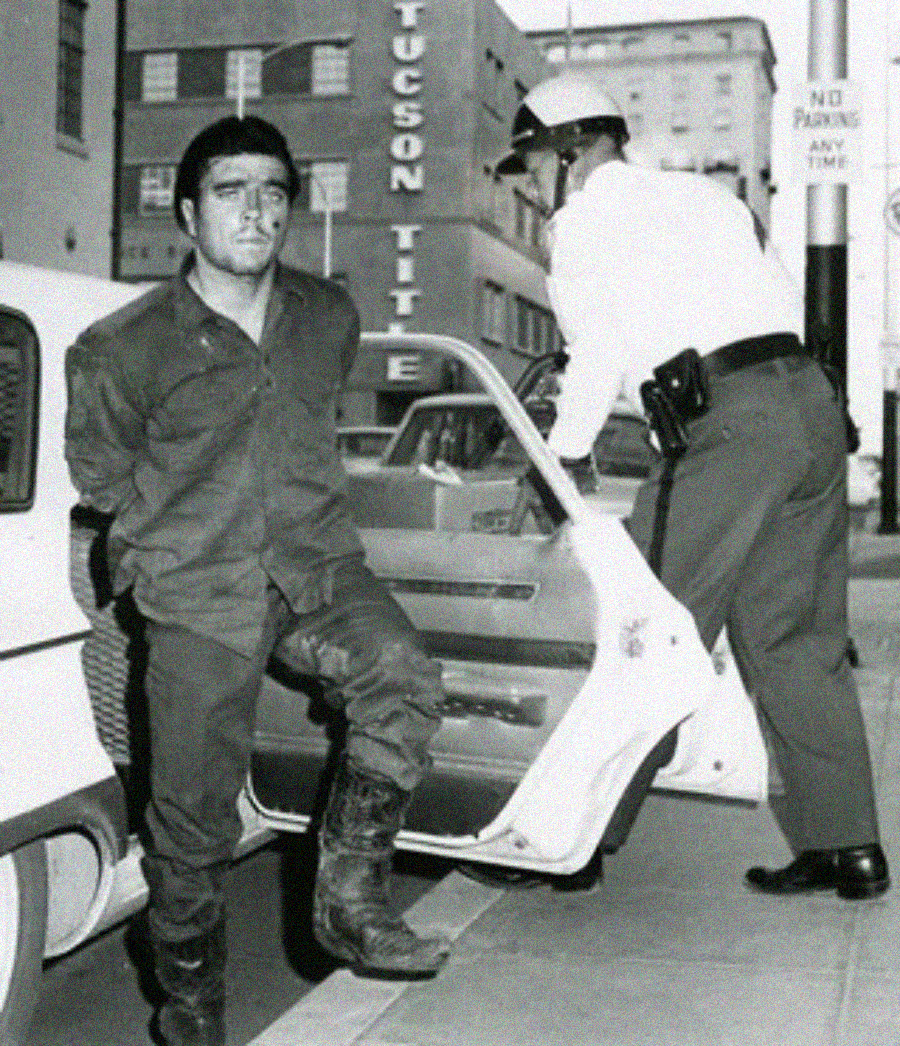A man in rugged clothing and boots is being escorted by a police officer to a car on a city street. The officer holds the car door open while the man appears to have his hands behind his back.