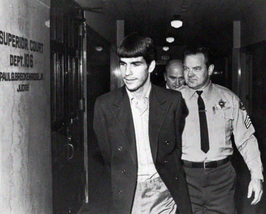 A young man in a suit, handcuffed and escorted by two uniformed officers, walks down a courthouse hallway with a sign reading "Superior Court Dept. 106" visible on the wall.