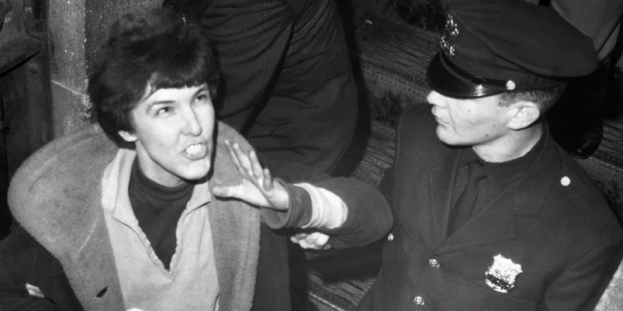 A police officer in uniform extends his hand to stop or restrain a woman, who appears to be speaking or shouting, on a staircase. The scene is in black and white and suggests a tense interaction.
