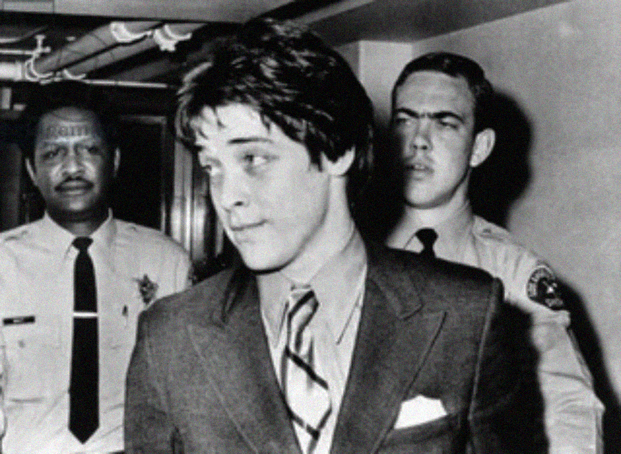 A young man in a suit and tie is escorted by two uniformed officers down a hallway. The man looks to the side with a neutral expression while the officers walk closely behind him. The photo is in black and white.