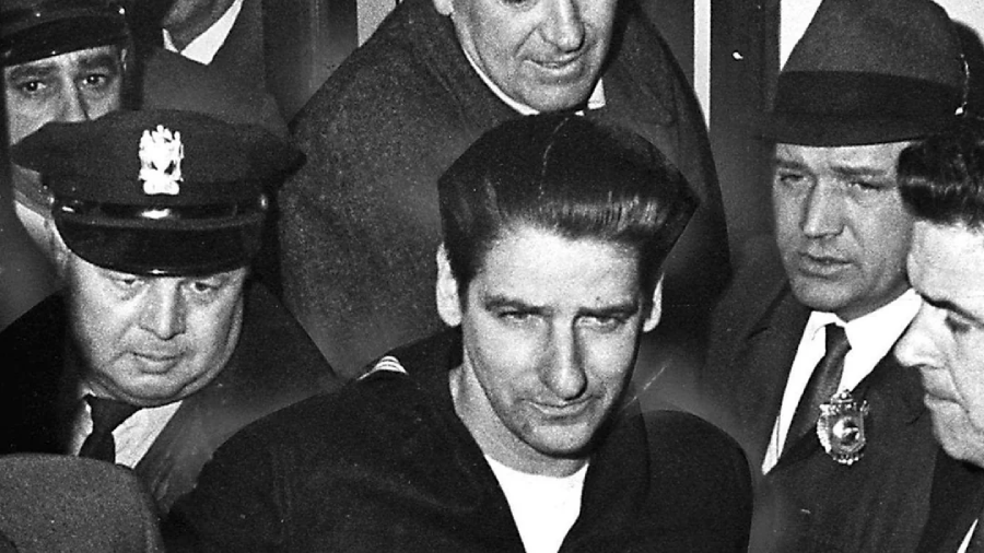 A black-and-white photo shows a man surrounded by police officers and officials, some wearing uniforms and hats, as they escort him through a crowd. The expressions appear serious and tense.