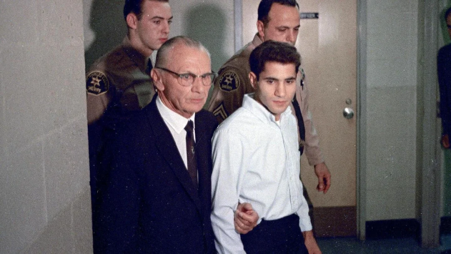 A young man in a white shirt is escorted by an older man in a suit and two uniformed officers down a hallway with beige walls. The group appears to be moving through a building.