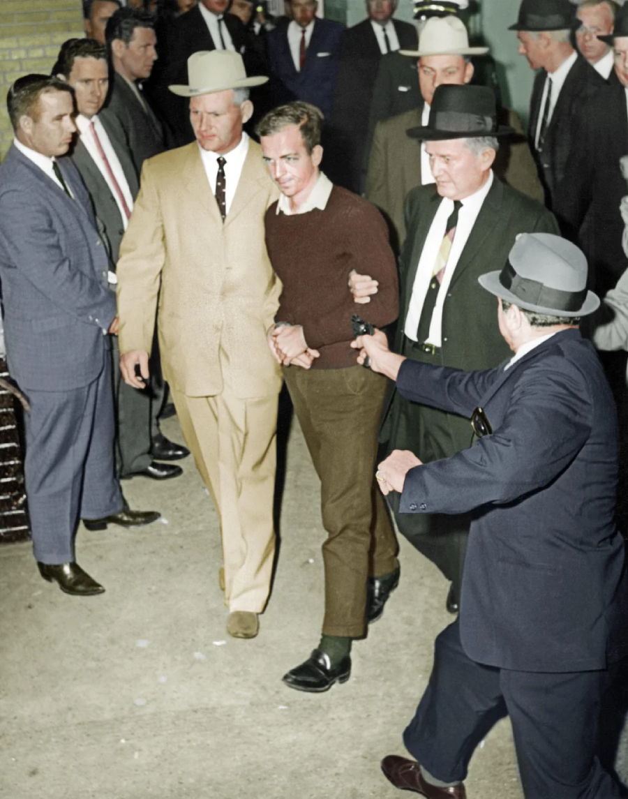 A young man in a brown sweater, handcuffed, is escorted by several men in suits and hats as a crowd looks on. One man in a blue suit points a gun at the young man. The scene appears tense and crowded.
