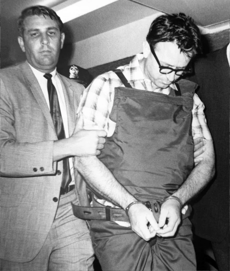 A handcuffed man wearing glasses and a protective vest is escorted by a police officer in a suit. The man looks down while the officer firmly holds his arm.