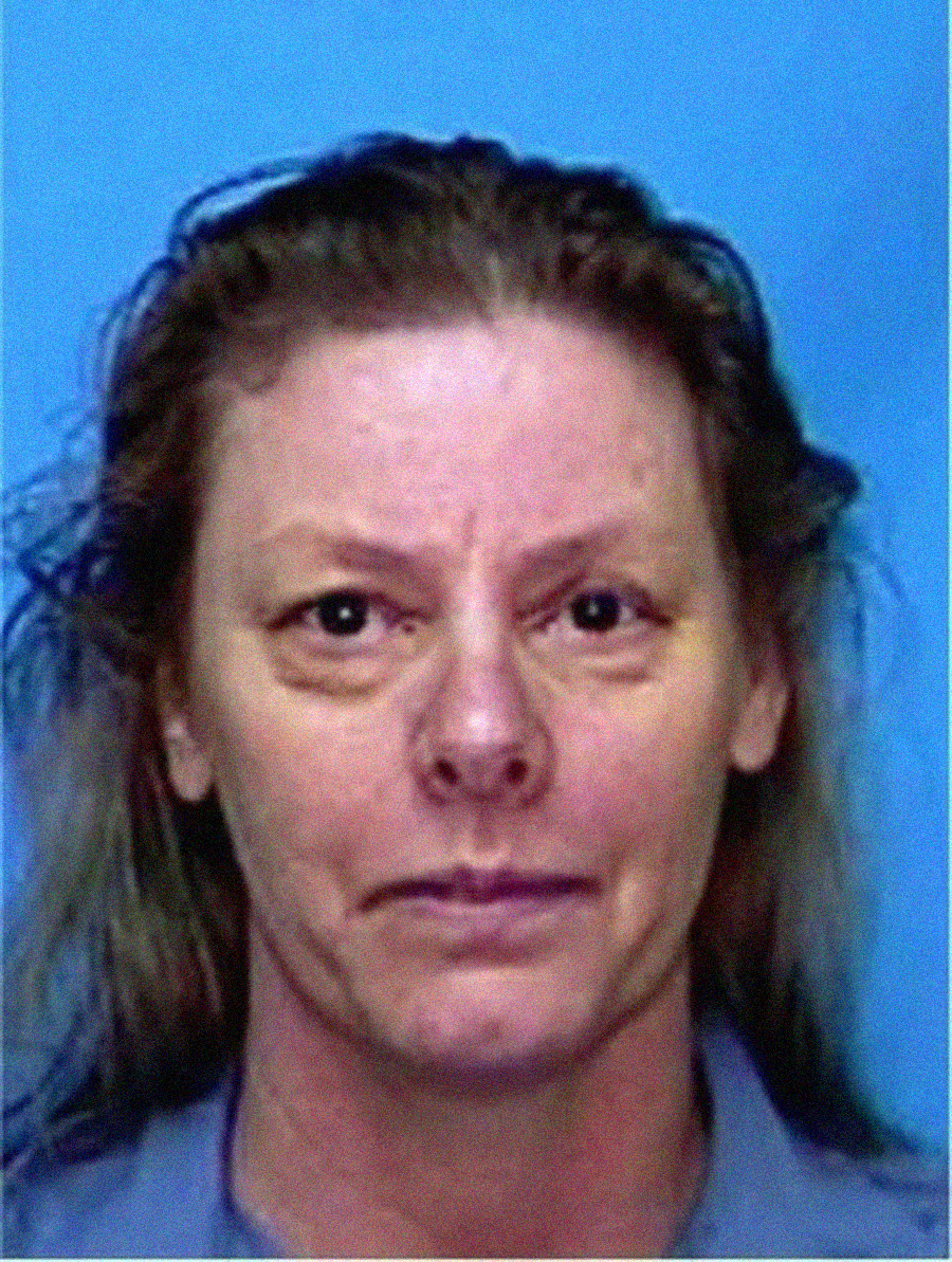 A woman with light skin and shoulder-length brown hair is shown against a blue background. She has a neutral expression and is wearing a gray shirt.