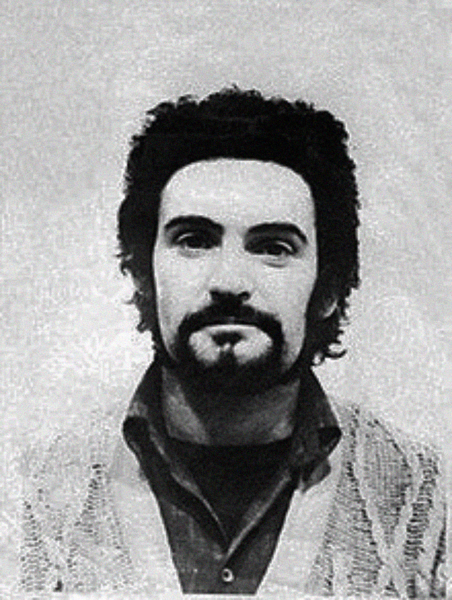 A black and white portrait of a man with dark, curly hair, a full beard, and mustache, wearing a collared shirt and a textured sweater, looking straight at the camera with a neutral expression.