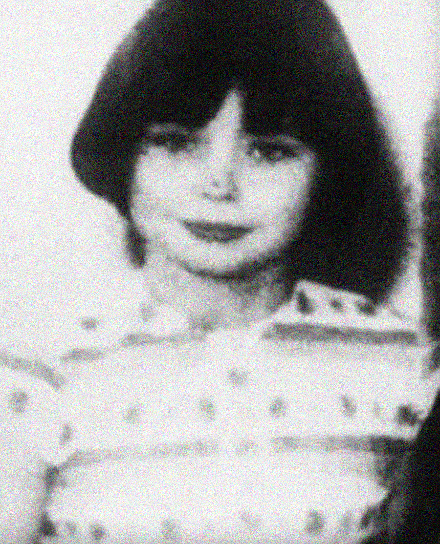 A grainy, black-and-white photograph of a young child with dark, chin-length hair and bangs, wearing a patterned shirt and looking at the camera with a slight smile.