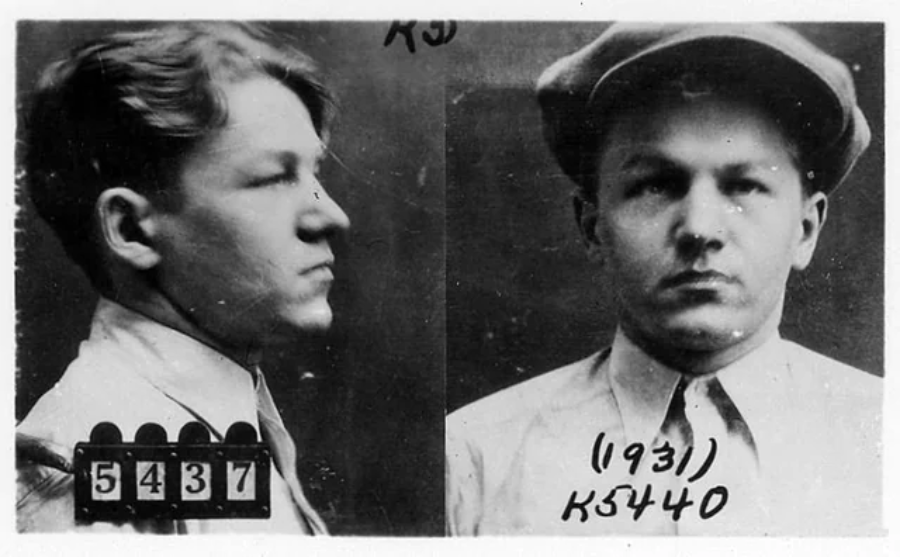 Black and white mugshot of a young man shown in profile and front view. He has short hair, wears a collared shirt and cap, and holds black placards with identification numbers.