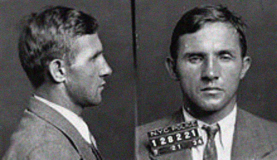 Black and white mugshot of a man in a suit and tie, shown from the side and front. He has short hair and holds a placard with numbers and letters.