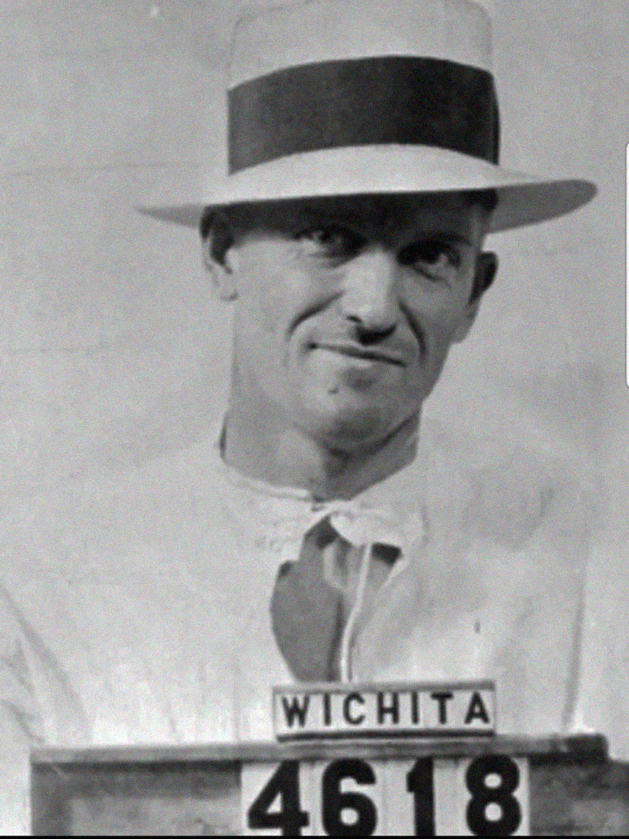 Black-and-white mugshot of a man wearing a light-colored shirt and a wide-brimmed hat with a dark band. He is holding a sign that reads "Wichita 4618" and looking slightly to the side.