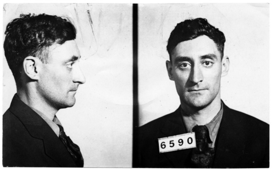 Black and white mugshot of a man with dark hair, shown in both profile and frontal views. He is wearing a suit and has a tag with the number 6590 around his neck.